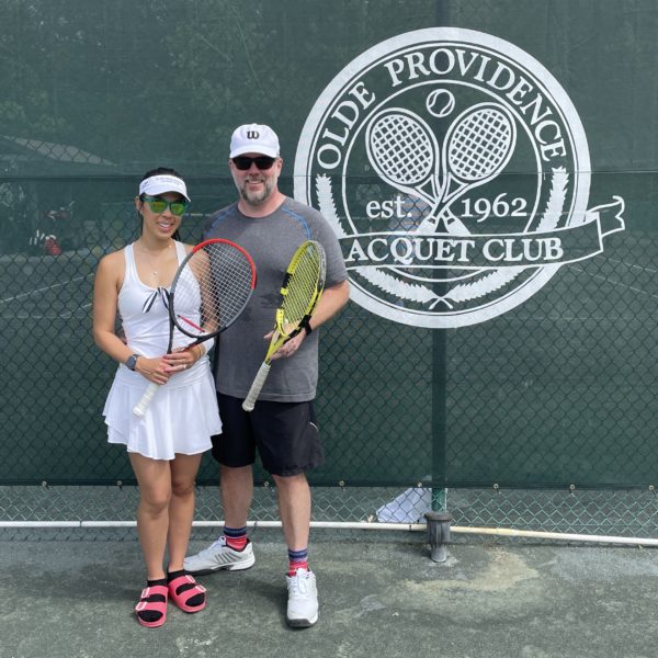 Caroleen and Joe are standing with their tennis rackets in front of the Olde Providence Racquet Club logo at the tennis court